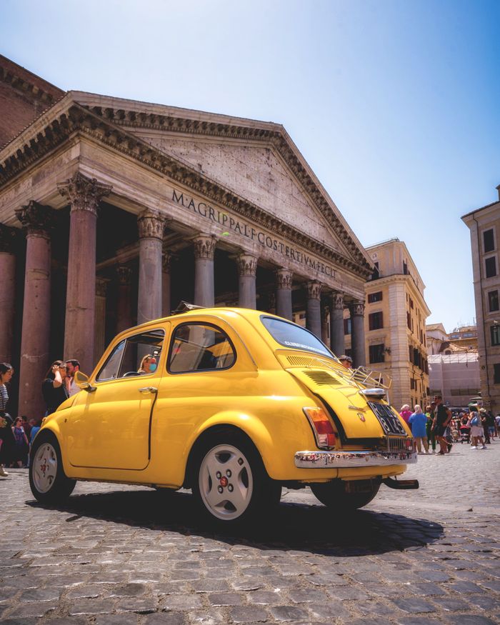 A classic yellow Fiat 500 parked outside the historic Pantheon in Rome on a sunny day.
