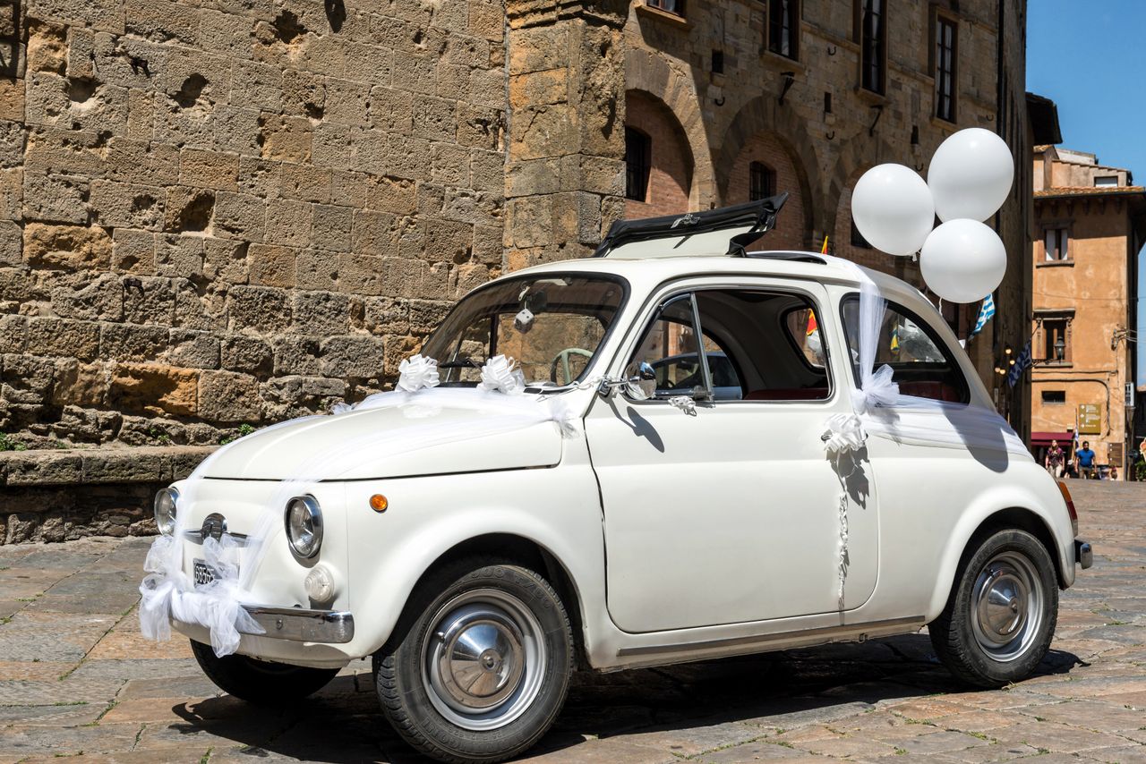 A classic Fiat 500 adorned with wedding decorations and white balloons on a sunny street in Volterra, Italy.