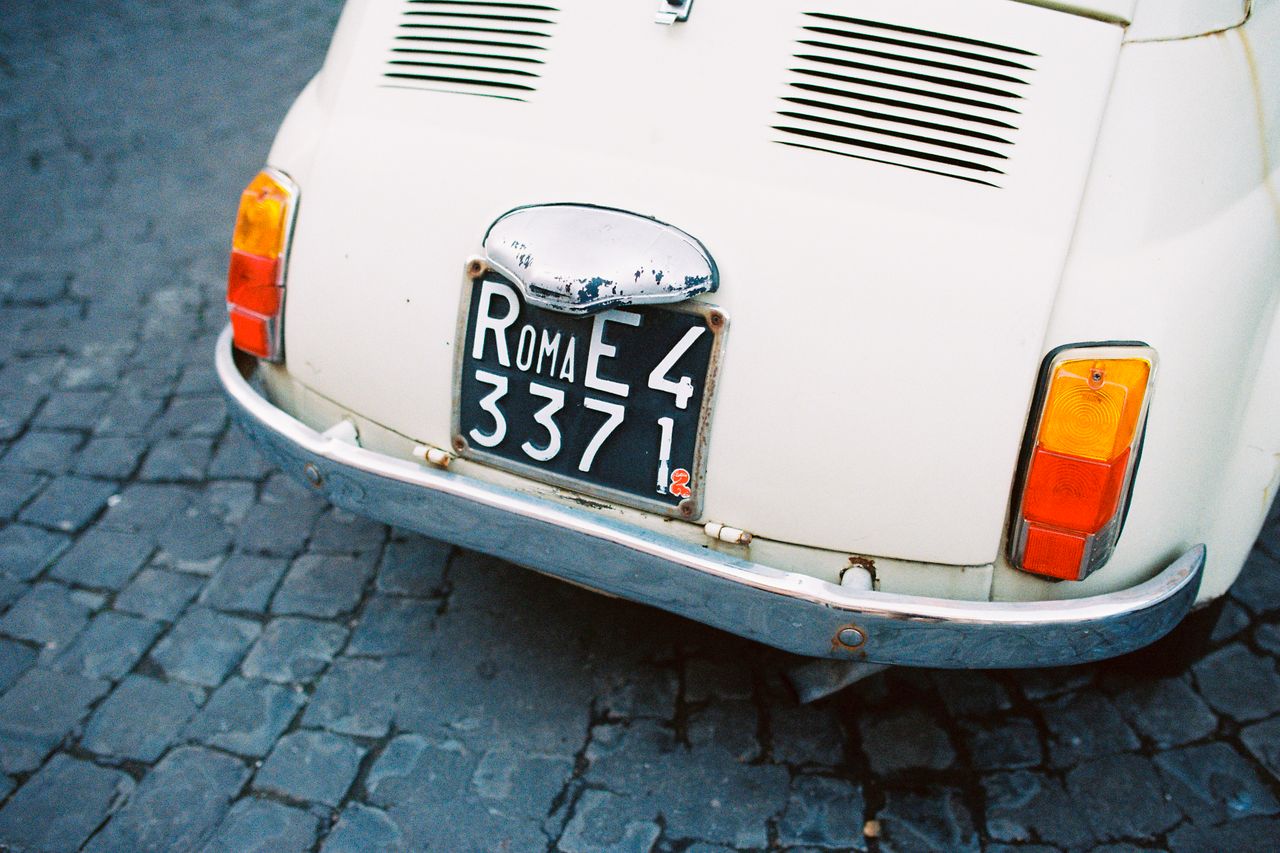 Close-up of a classic Fiat 500's bumper with a Roman license plate on cobblestone streets.